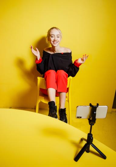 Young fashion girl blogger dressed in red trousers and black jacket takes a selfie on the smartphone standing possing sitting on the stool in the room with yellow walls and furniture .